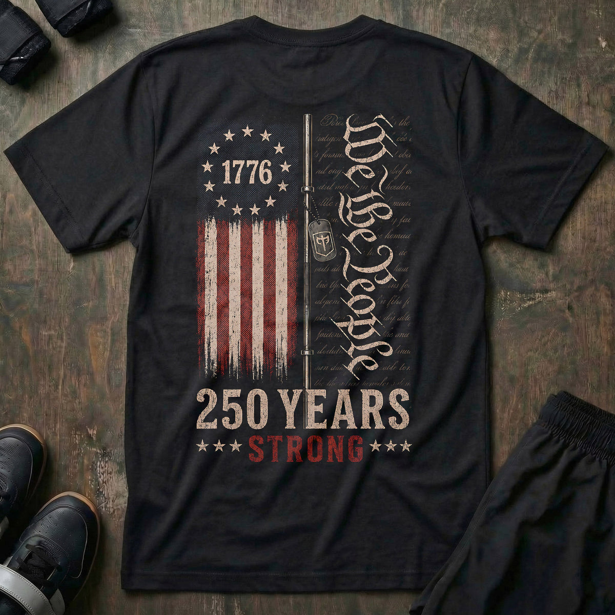 250 Years Strong tee in Black: a distressed 1776 flag split alongside a barbell with a CB dog tag hanging from it