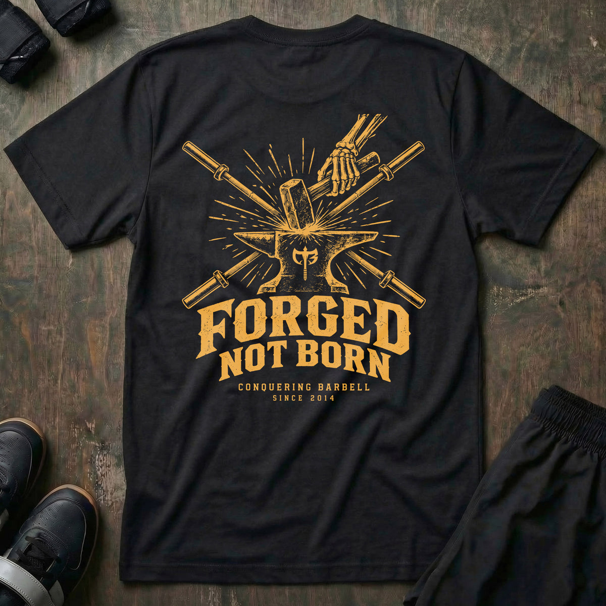 Forged, Not Born tee in Black: a skeletal hand driving a hammer down onto an anvil