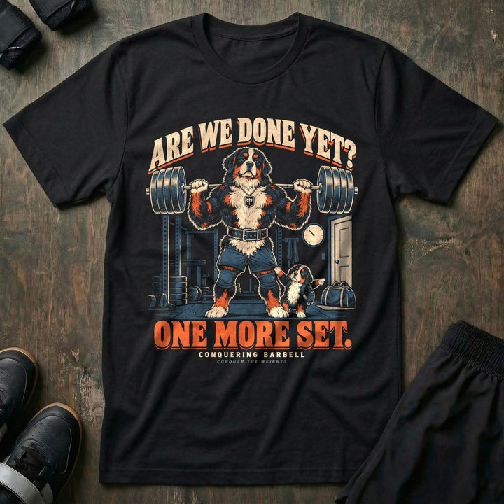 Are We Done Yet? One More Set - Tee front graphic in Black with a Bernese mountain dog dad with a loaded barbell on his back while a puppy tugs his shorts and points at the door - Conquering Barbell