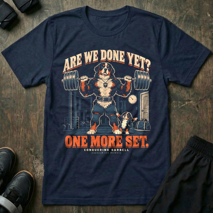 Are We Done Yet? One More Set - Tee front graphic in Navy with a Bernese mountain dog dad with a loaded barbell on his back while a puppy tugs his shorts and points at the door - Conquering Barbell