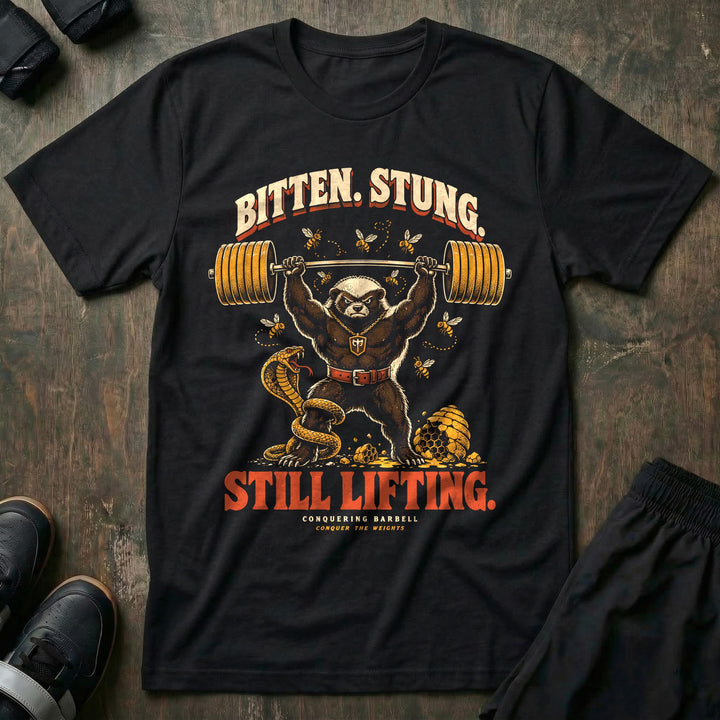 Bitten Stung Still Lifting - Tee front graphic in Black with a honey badger pressing a barbell overhead while bees swarm and a cobra coils - Conquering Barbell