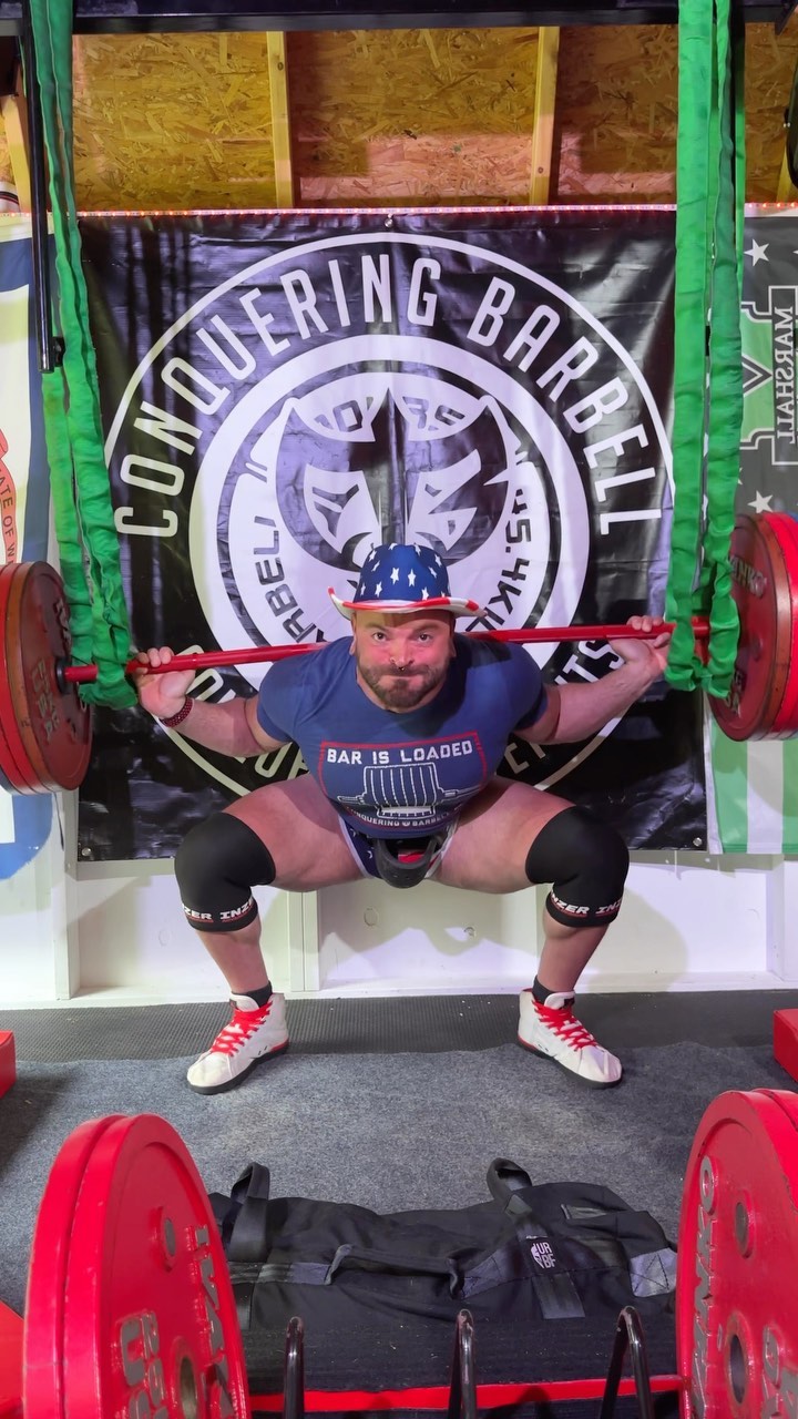 @paynterperiodization squatting in the Bar Is Loaded tee in front of a Conquering Barbell flag