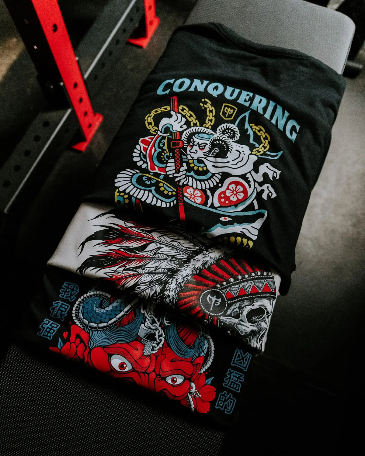 Three Conquering Barbell tees folded on a gym bench next to a red power rack