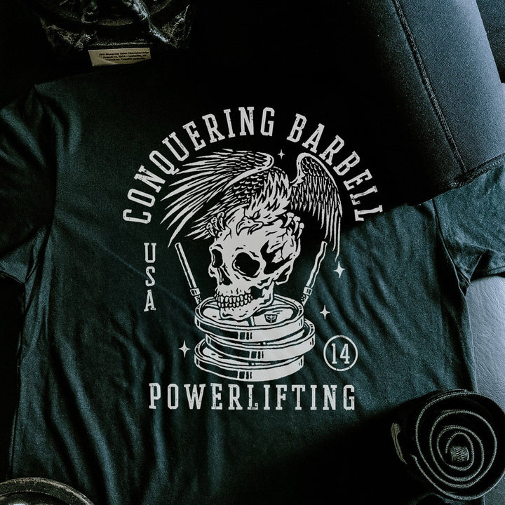 Conquering Barbell Eagle tee in Black: an eagle with spread wings perched on a skull atop stacked plates