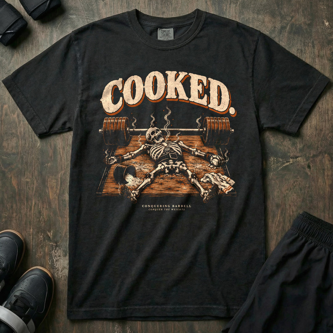Cooked - Relaxed Fit Tee front graphic in Black with a smoking skeleton laid out flat on the platform beside a loaded barbell - Conquering Barbell