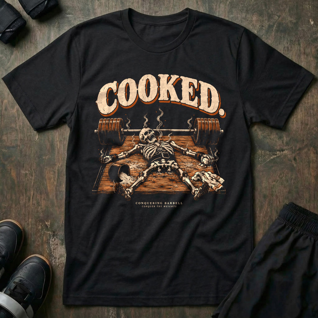 Cooked - Tee front graphic in Black with a smoking skeleton laid out flat on the platform beside a loaded barbell - Conquering Barbell