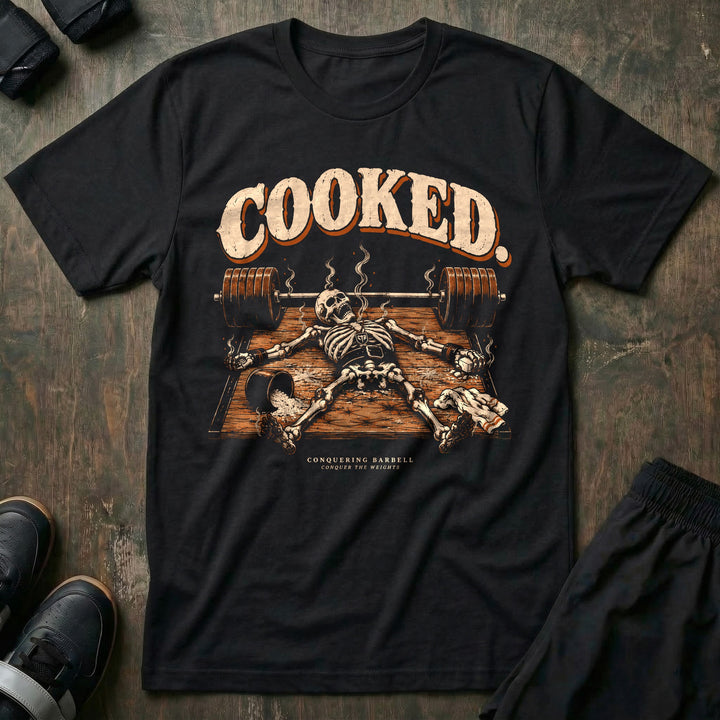 Cooked - Tee front graphic in Black with a smoking skeleton laid out flat on the platform beside a loaded barbell - Conquering Barbell