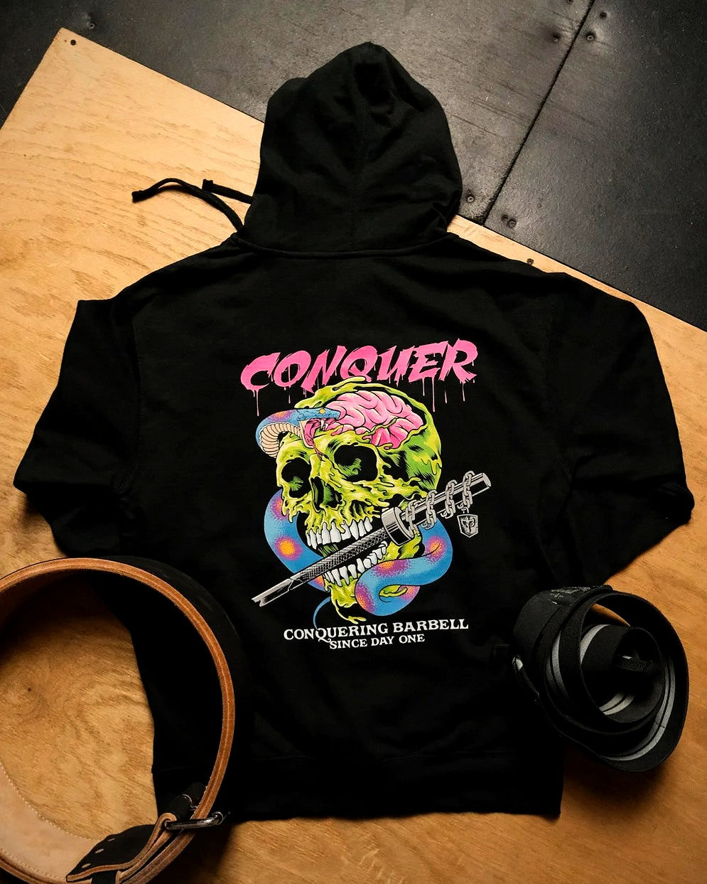 Conquering Barbell pullover hoodie in black with a full-color back print, on a lifting platform