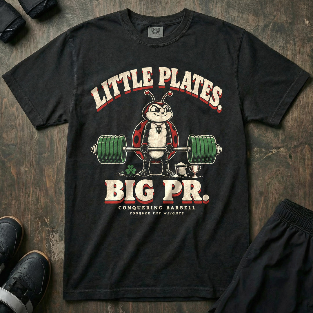 Little Plates Big PR - Relaxed Fit Tee front graphic in Black with a ladybug deadlifting a loaded barbell beside a tiny trophy and a four-leaf clover - Conquering Barbell