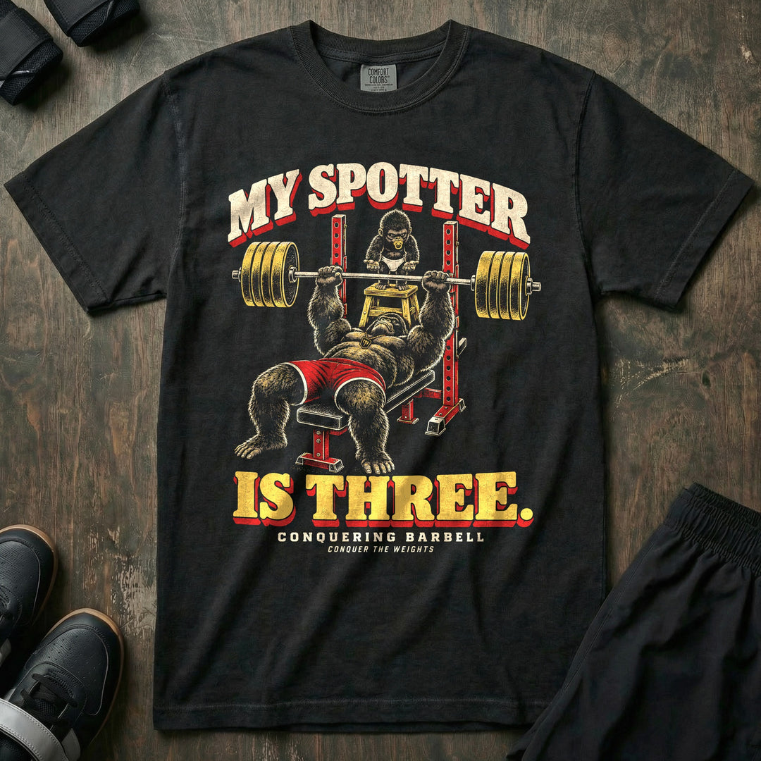 My Spotter Is Three - Relaxed Fit Tee front graphic in Black with a gorilla dad bench pressing a loaded barbell while a baby gorilla in a diaper spots him from a step stool - Conquering Barbell