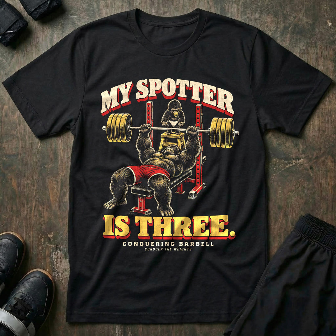 My Spotter Is Three - Tee front graphic in Black with a gorilla dad bench pressing a loaded barbell while a baby gorilla in a diaper spots him from a step stool - Conquering Barbell