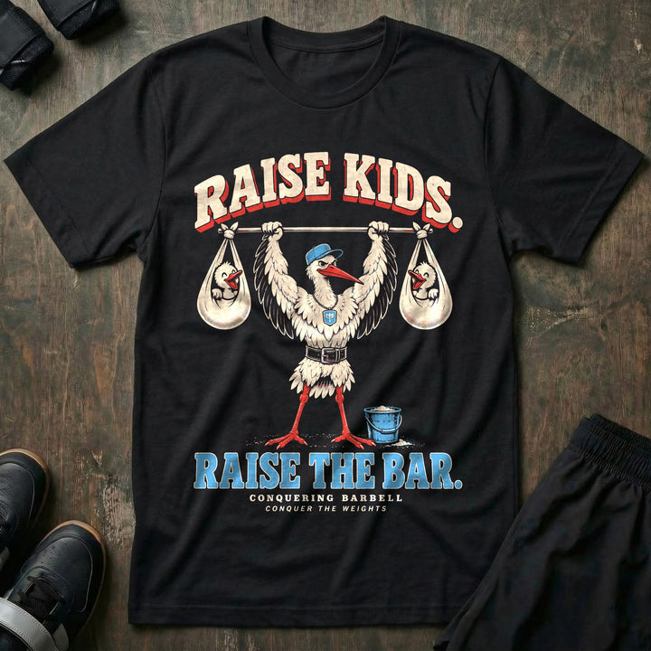 Raise Kids. Raise the Bar - Tee front graphic in Black with a stork dad in a ball cap pressing a barbell overhead with a baby bundle hanging from each end - Conquering Barbell