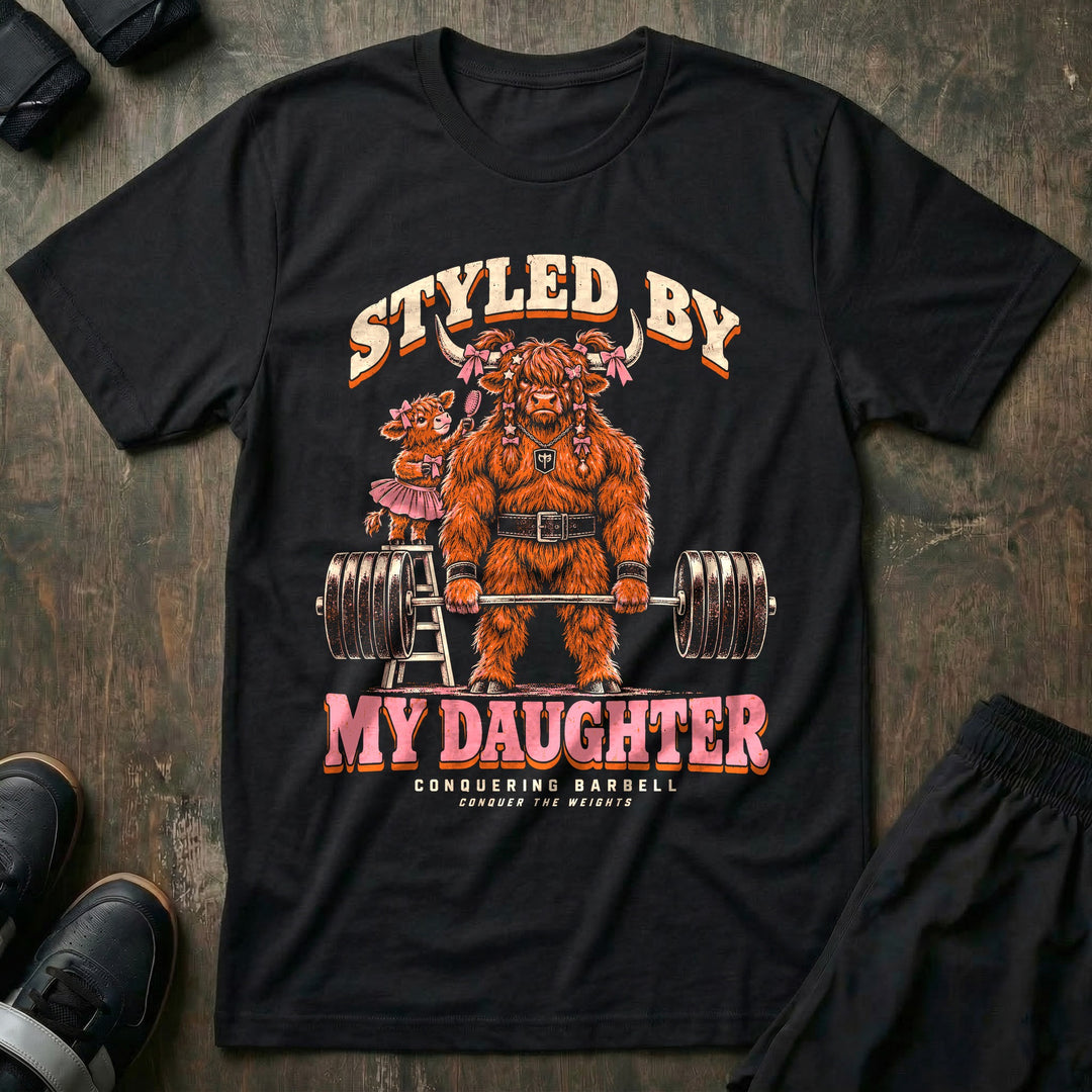 Styled by My Daughter - Tee front graphic in Black with a Highland bull dad with pink bows in his hair deadlifting a loaded barbell while his daughter in a tutu brushes his hair from a ladder - Conquering Barbell
