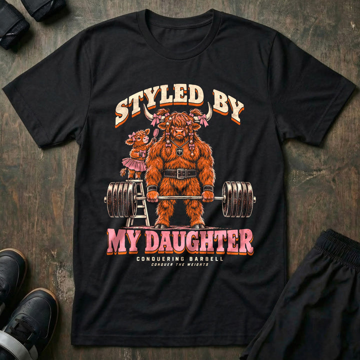 Styled by My Daughter - Tee front graphic in Black with a Highland bull dad with pink bows in his hair deadlifting a loaded barbell while his daughter in a tutu brushes his hair from a ladder - Conquering Barbell