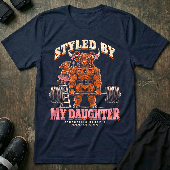 Styled by My Daughter - Tee front graphic in Navy with a Highland bull dad with pink bows in his hair deadlifting a loaded barbell while his daughter in a tutu brushes his hair from a ladder - Conquering Barbell