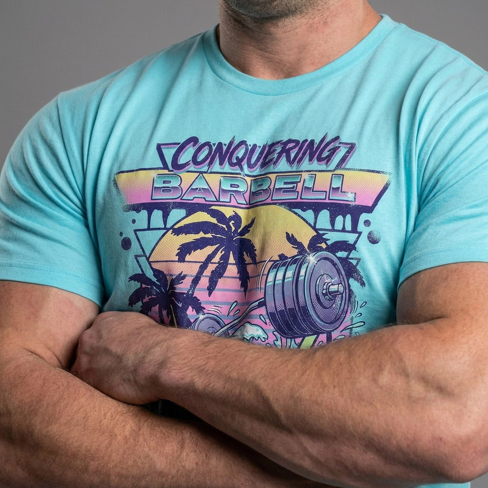 Suns Out Guns Out tee in Tahiti: a barbell rising from ocean waves with palm trees silhouetted against a neon sunset