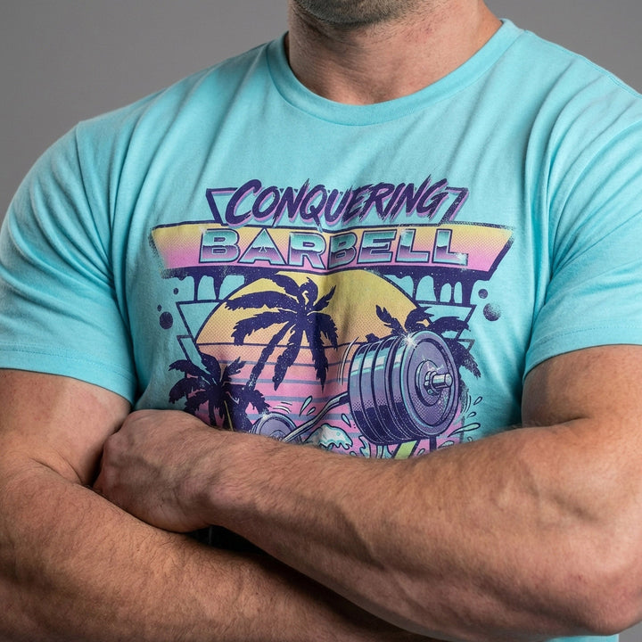 Suns Out Guns Out tee in Tahiti: a barbell rising from ocean waves with palm trees silhouetted against a neon sunset