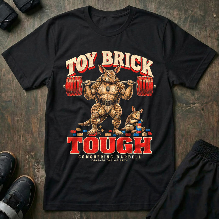 Toy Brick Tough - Tee front graphic in Black with an armadillo dad with a loaded barbell on his back stepping on a toy brick while a young armadillo stacks building blocks - Conquering Barbell