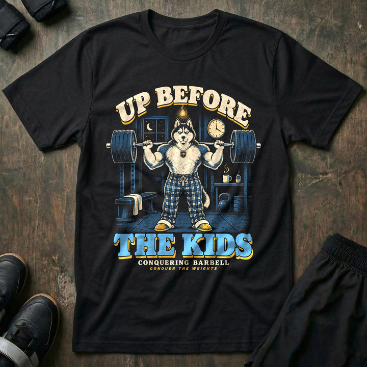 Up Before the Kids - Tee front graphic in Black with a husky dad in plaid pajama pants squatting a loaded barbell in a garage gym at night with a coffee mug and baby monitor on a shelf - Conquering Barbell