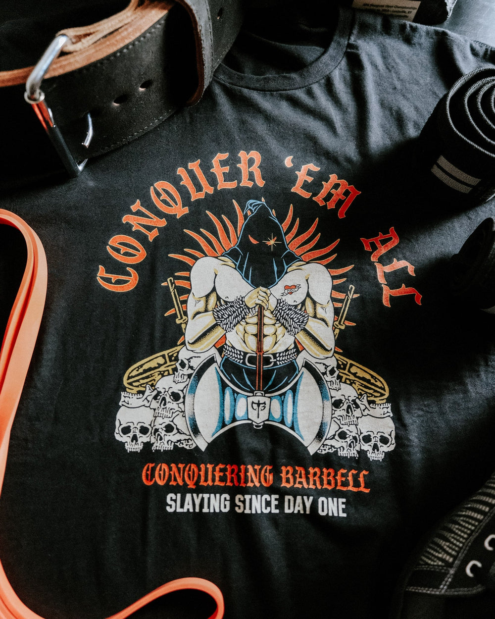 Conquer 'Em All tee in Black: a hooded executioner enthroned on a pile of skulls and loaded barbells