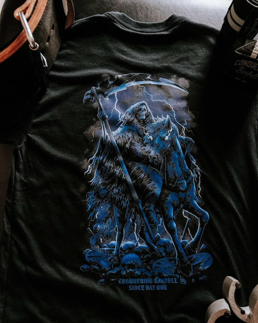 The Reaper tee in Black: a hooded reaper with his scythe riding out of the shadows in cold grey and steel blue